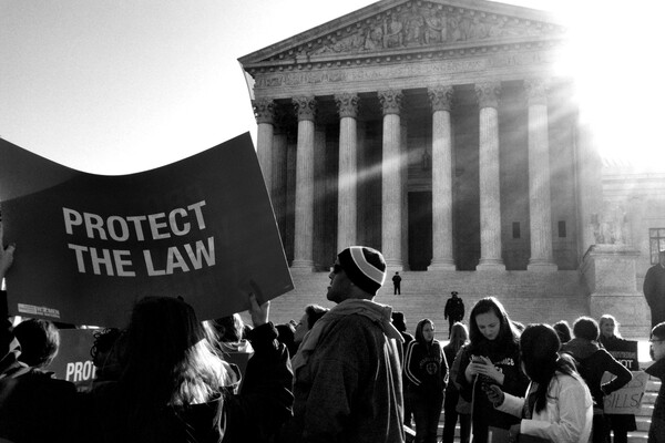 Group of people outside the steps of the Supreme Court, one person holds a large sign that reads PROTECT THE LAW.