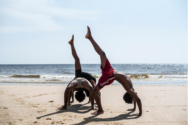 Three dancers doing back bands with one leg raised on a sunlit beach. 