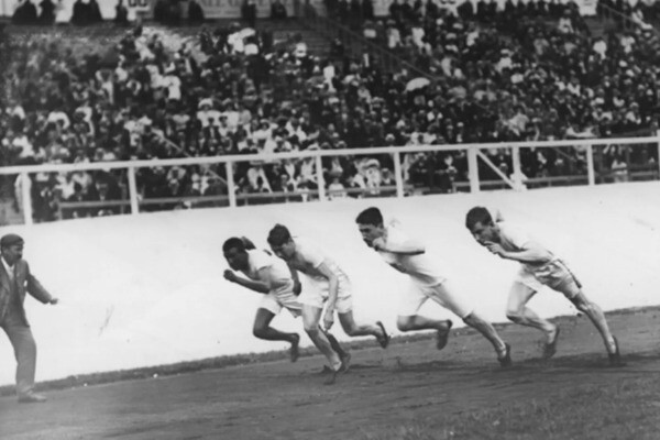 John Baxter Taylor Jr., left, races three other competitors at the 1908 Summer Olympics in London. All four runners are wearing white shirts and shorts.