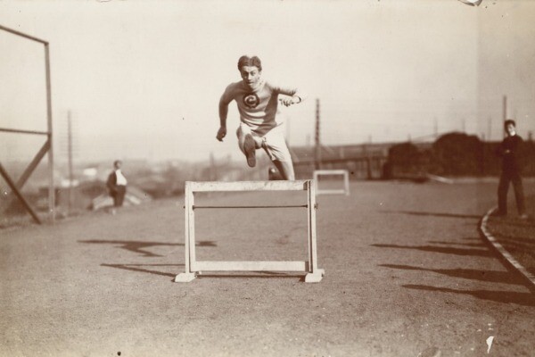 Circa 1900, Alvin Christian Kraenzlein jumps over a hurdle with two observers watching in the background.
