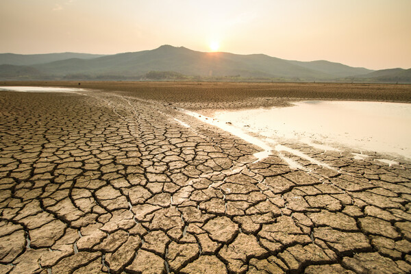 A dried up desert with a small amount of water on the right-hand side. The sun is blazing in the background, in front of mountains.