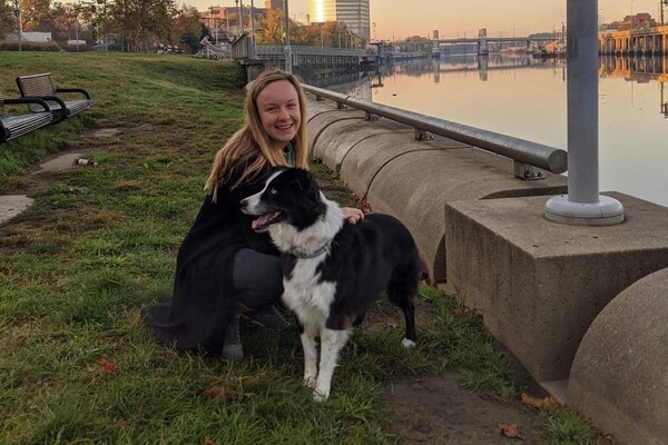 Smiling person kneels with dog along riverbank