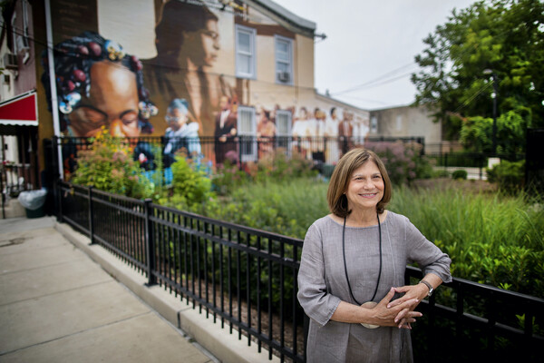 A person standing along a black iron fence, one arm hanging over the fence. In the background are trees and a blurred out mural.