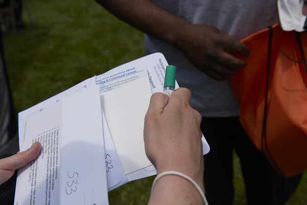 A pair of hands holding a cancer screening kit vial in one hand and paperwork in another extended to a person standing in a park.
