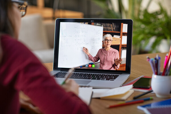 In the foreground, a blurred out student holding a pencil over a notebook watching a math lesson on a computer screen. In the background are blurred out plants, table and chairs.