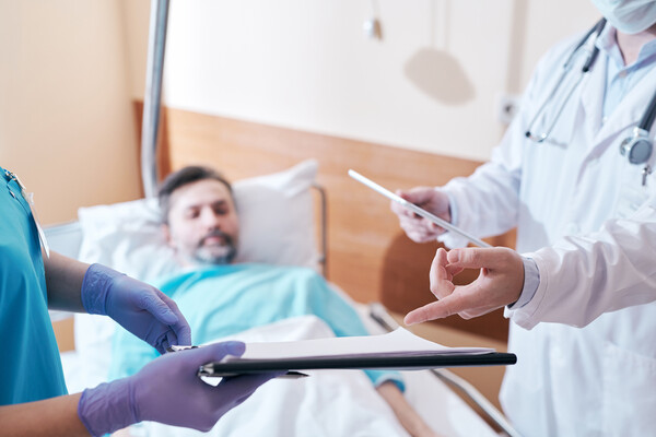 Patient laying in hospital bed while a doctor and a nurse compare notes regarding the patient’s medical chart.