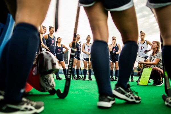 During practice at Vagelos Field, Head Coach Colleen Fink, kneeling, addresses her players, who are standing.