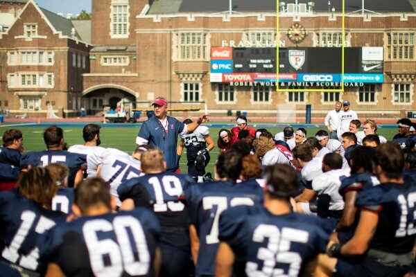 Head Coach Ray Priore, who is standing, addresses the football team, who are kneeling in a semicircle on Franklin Field.
