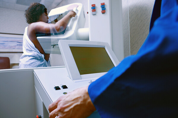 A Black woman standing at a mammogram machine with a mammogram technician in the foreground.