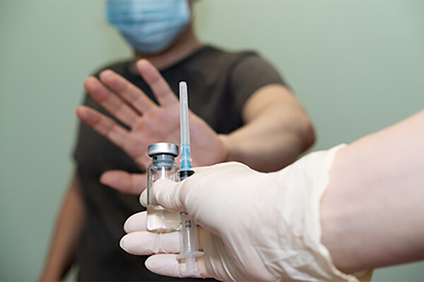 Masked person holding up hand in defense against a vial of vaccine being offered by a gloved hand.