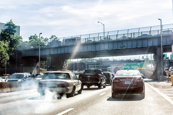 vehicles in traffic on a highway in daylight, one older car has exhaust coming out of its tailpipe.