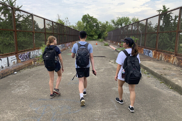 Three students with backpacks walk across a foot bridge with high fences and graffiti on the low walls in Philadelphia.