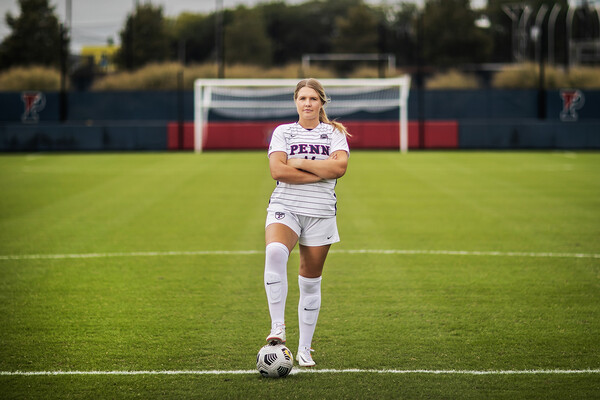 Wearing her white Penn jersey and white shin guards, Mia Shenk stands with her arms folded and with her right foot on a soccer ball at Rhodes Field.