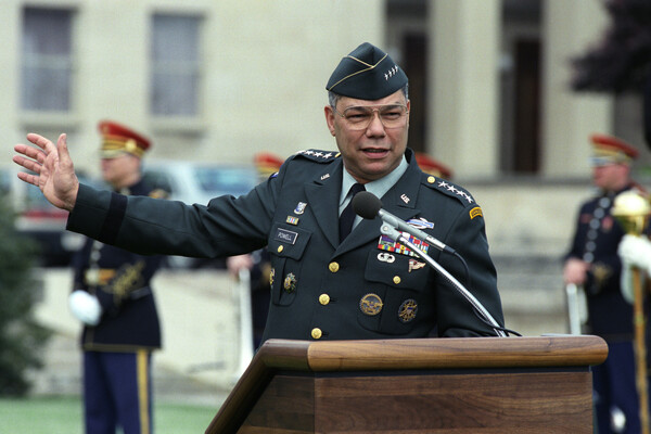 Colin Powell, wearing full military garb and standing at a brown wooden podium with a microphone, gestures to his right, sweeping his arm that direction
