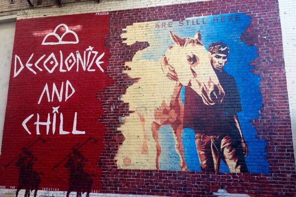 A mural on a brick wall. On the left-hand side, on a red background, are the word "Decolonize and Chill" in white. Beneath that are the silhouette of two men riding horses. On the right-hand side is a person with a horse, beneath the words "We are still here."