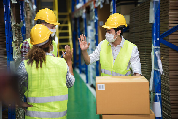 Three workers in a stock room wearing safety vests, hard hats, and face masks, wave hello to each other.