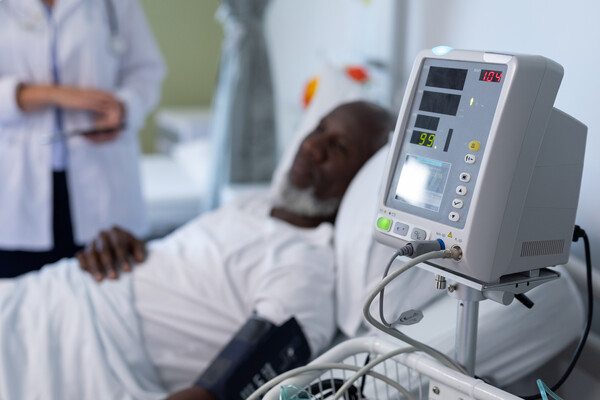 African American person laying in a hospital bed with a blood pressure monitor, a medical professional in a white coat stands beside the bed.