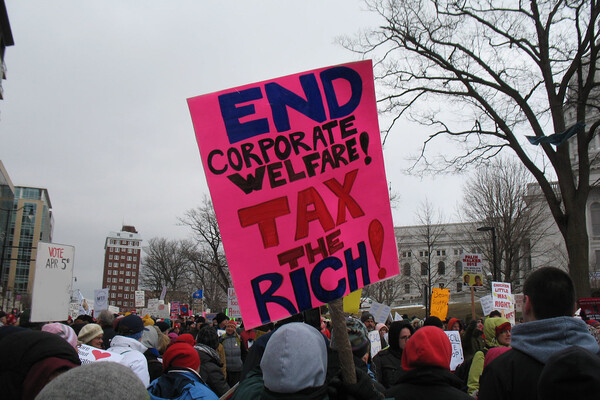 protesters are seen from behind on a city street, one holding a hot pink sign with handlettering reading "end corporate welfare! tax the rich!