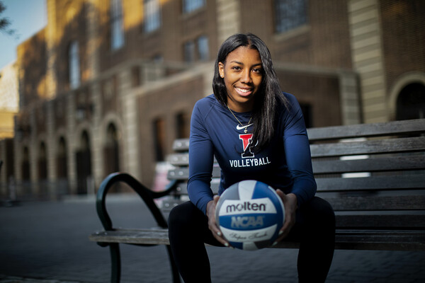 Autumn Leak, holding a blue and white volleyball, sits on a bench outside of the Palestra. She is wearing a blue Penn volleyball shirt.