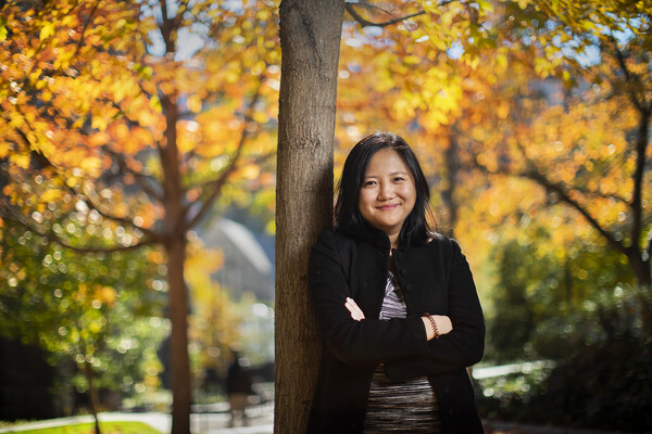 Woman leans against a tree with her arms crossed, looking into the camera, with other trees in fall colors behind her on a sunny day