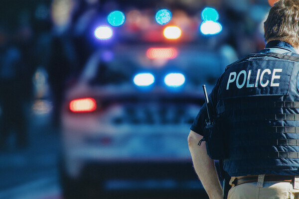 A person wearing a "POLICE" vest and a walkie talkie, standing in front of a blurry police car with its lights flashing.