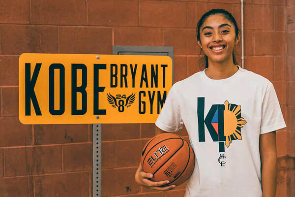 Kayla Padilla smiles while holding a basketball in a t-shirt she designed in front of a sign that reads Kobe Bryant Gym.