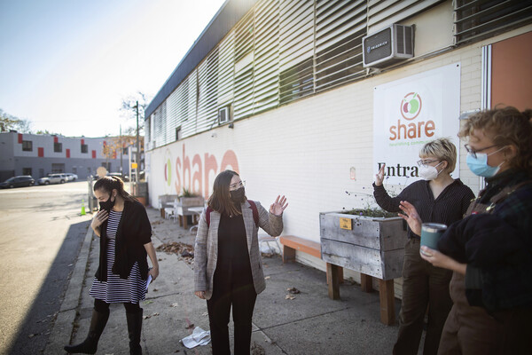 Four women in front of the Share food bank, and the two in the middle are waving to each other