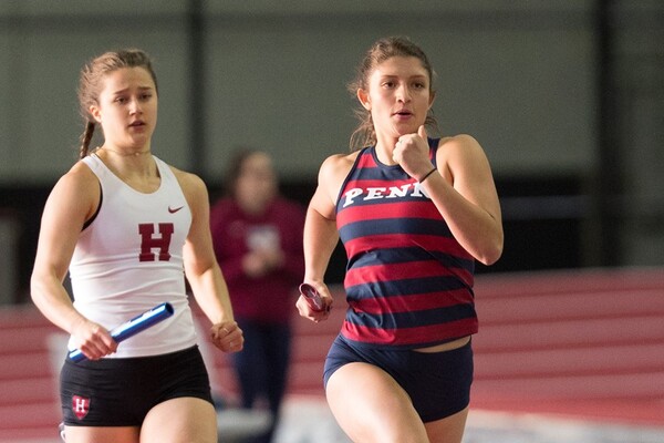 Haley Rizek runs around an indoor track with a baton in her hand, passing by a runner from Harvard.