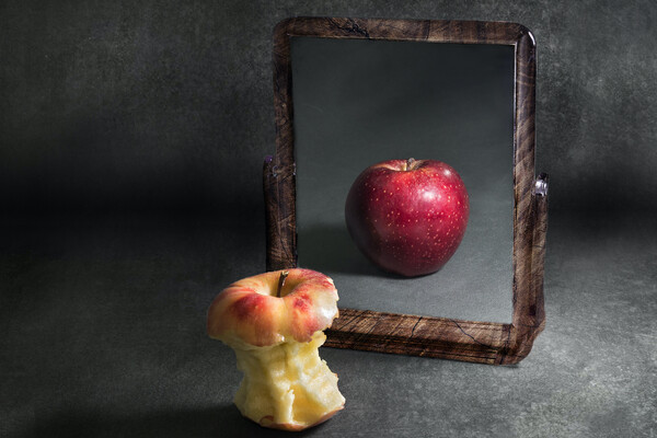 A mostly eaten apple in front of a mirror showing a whole, uneaten apple.