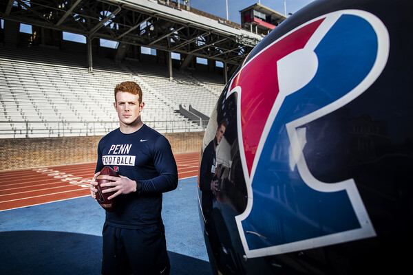 Standing on Franklin Field wearing a Penn football shirt, Brian O'Neill holds a football with both hands. A Penn football helmet sits at right.