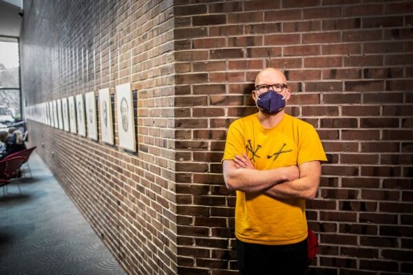 Mark Stockton leans against brick wall on corner of wall of portraits