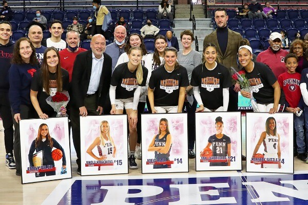 Seniors on the women's basketball team stand at midcourt at the Palestra with posters of them in front. Family and friends are behind them.