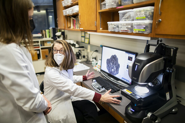 Kathy Morrison sitting at a computer in a lab, gesturing to grad student Moriah McKenna. They both wear white lab coats. In front of them is a computer screen with what looks like a large rock. Next to the computer is a large microscope with its light on.