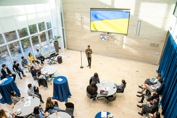 A person speaking into a microphone in front of people seated at tables at Perry World House with a digital Ukrainian flag on the wall.