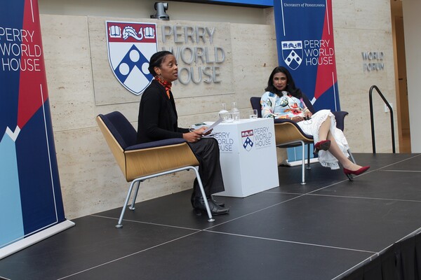 Two women sit on either side of a table onstage. The signage behind them reads, "Perry World House"