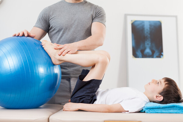 Young child laying on their back putting their feet on an exercise ball with an adult helping and an X ray on the wall.