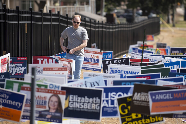 Man in sunglasses with earbuds walks throught a field covered in campaign posters