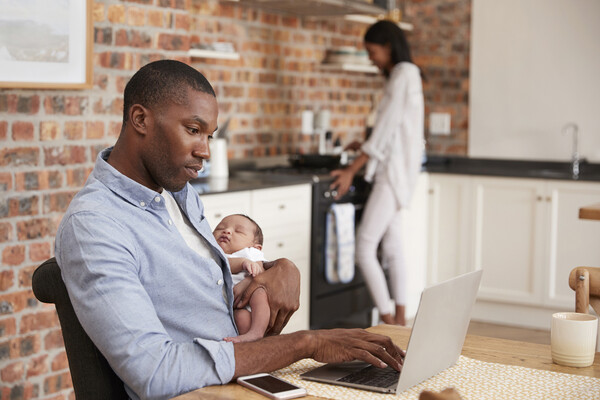 Parent siting at table holding a baby while working on laptop. Another person in the kitchen in the background.