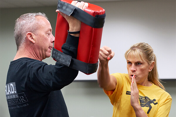 Deborah Millar demonstrates self-defense moves on a punching bag held by a person next to her.