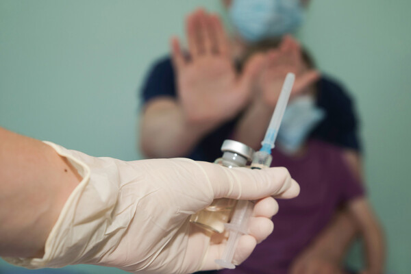 Gloved hand holding a vaccine vial and needle extended toward a parent holding their hands up to deny the shot.