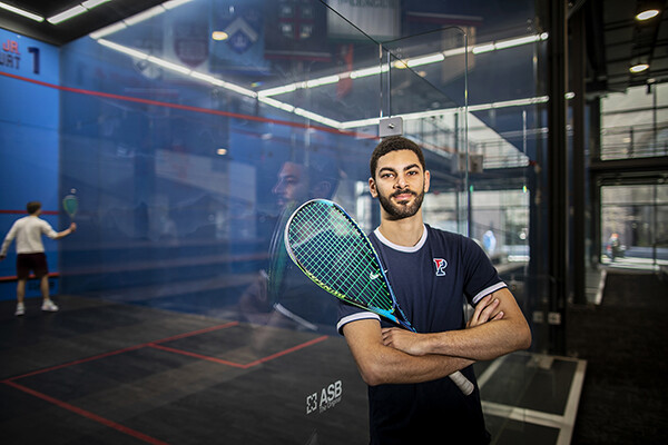 Aly Abou Eleinen, holding a racquet, stands inside the Penn Squash Center next to a glass court.