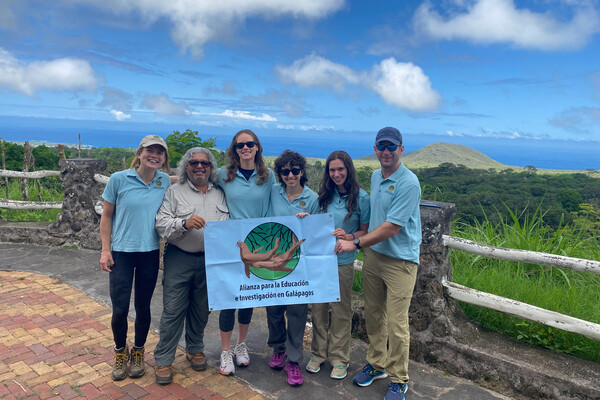 Group poses in a tropical marine landscape holding a sign that reads Allianza para la Educaion e Investigacion en Galapagos