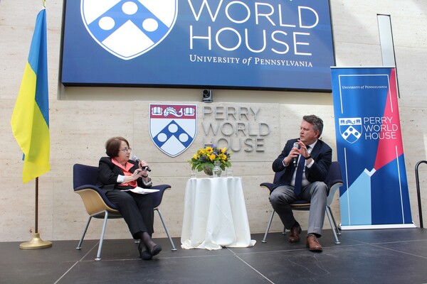 two people sitting on stage talking with Perry World House on the wall behind them and the Ukrainian flag beside them 
