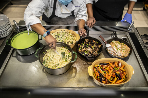 The arms of two people over an industrial-sized kitchen warmer, which holds a pot of green puree, two pans of pasta, a cast iron skillet of mushrooms, another cast iron skillet of fish with a spatula on top, and a bowl of multi-colored carrots. A stack of bowls sits off to the left of the image.