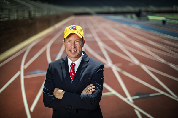 Steve Dolan, wearing a blue suit, red tie, and yellow hat, poses on the track at Franklin Field.