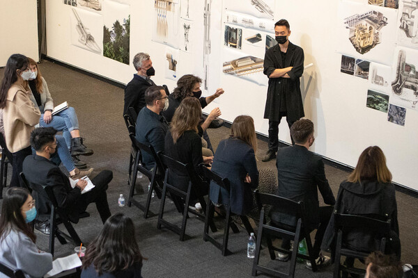 A person stands in front of a wall of photos and architectural renderings before a group of seated people.