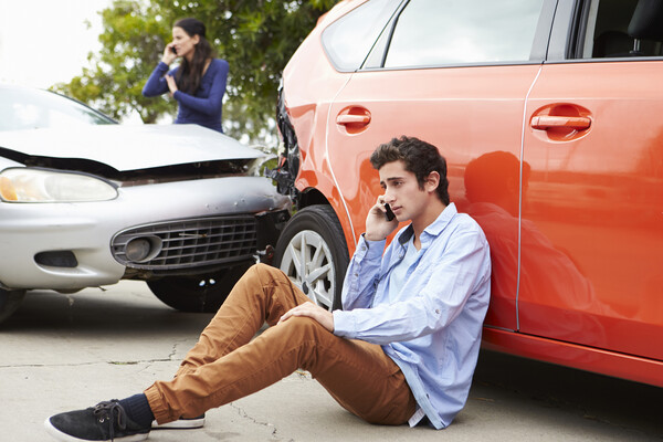 A teenager sits on the ground on a cell phone beside a vehicle that had crashed into another vehicle.