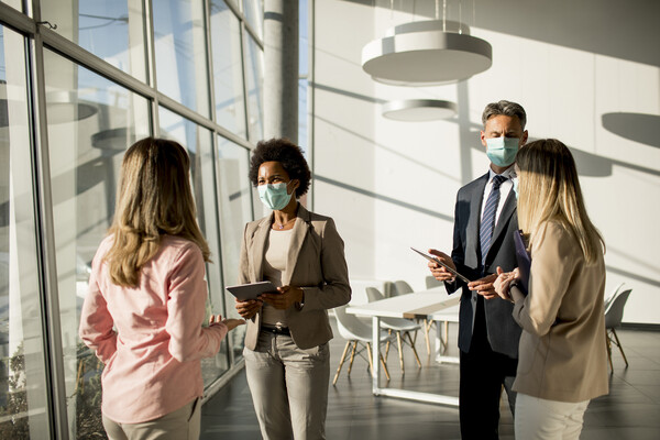 Four coworkers standing by sunlit windows having a conversation, wearing face masks.