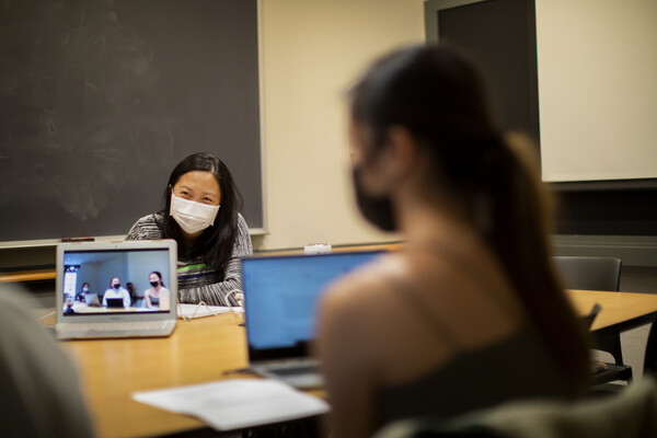 Penn history P.hD. candidate Sarah Yu sits at a table with a blackboard behind her, a laptop on the table showing a video of students