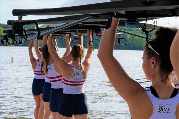 Members of the women's rowing team, wearing their white Penn jerseys, carry their boat into the water.